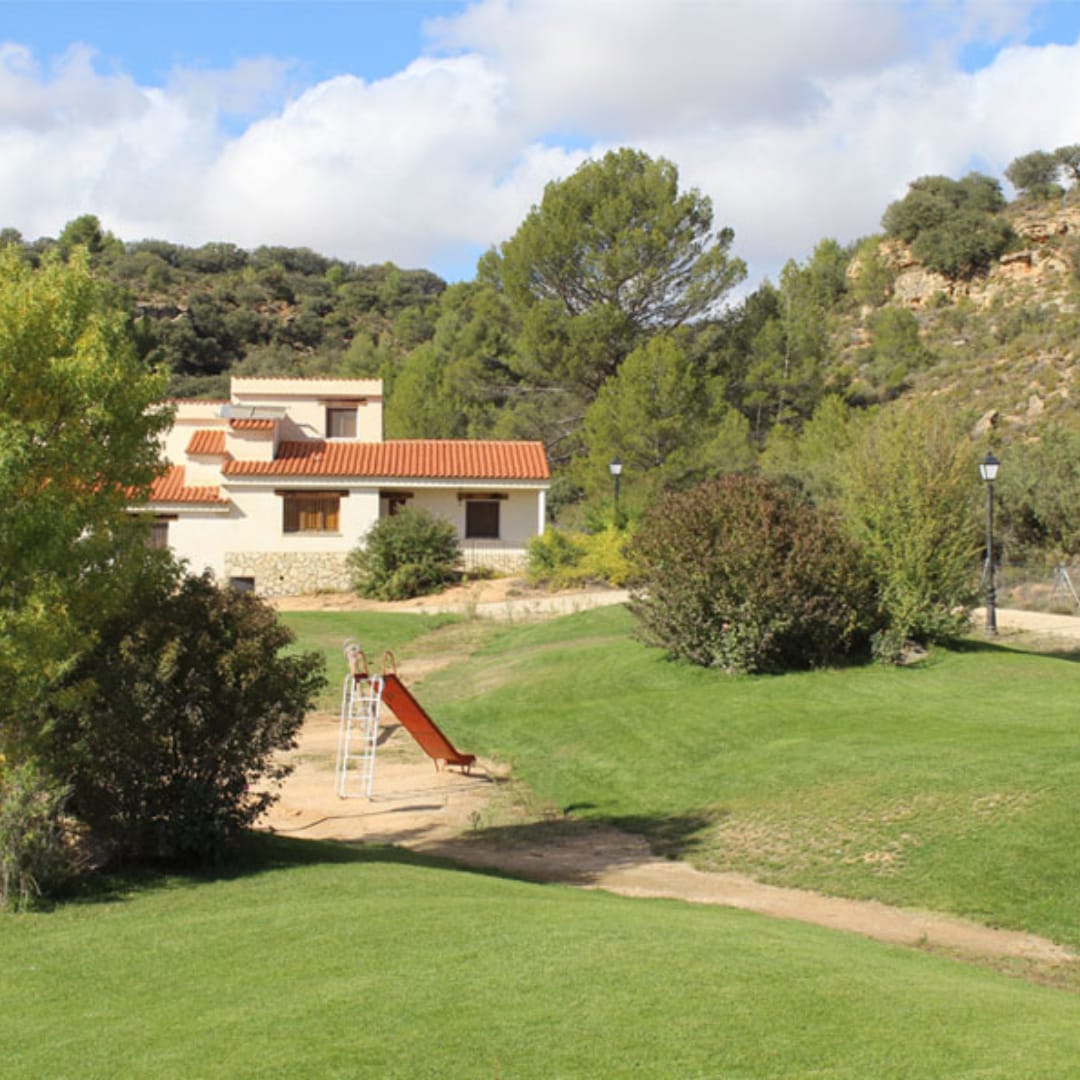 A spacious house is set amidst lush greenery, featuring a clay roof and multiple windows. A slide and play area are visible in the foreground, surrounded by well-maintained grass and shrubs. A natural landscape and low hills provide a serene backdrop.