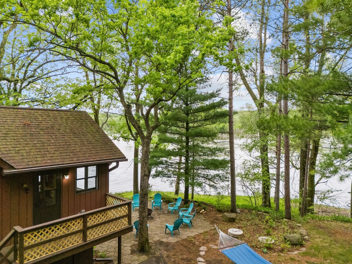 A serene lakeside view is presented, highlighting a cozy cabin surrounded by lush trees. Bright blue outdoor chairs are arranged in a circle, inviting relaxation. A hammock is visible nearby, enhancing the tranquil atmosphere. The peaceful waters of the lake can be seen in the background.