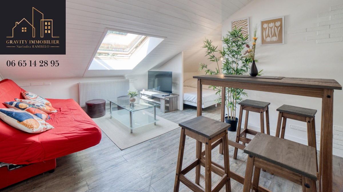 A bright studio space features a red sofa bed against the sloped ceiling, paired with a glass coffee table. A wooden dining table with four stools is positioned beside lush green plants, while a television stand occupies the corner, enhancing the functional living area.