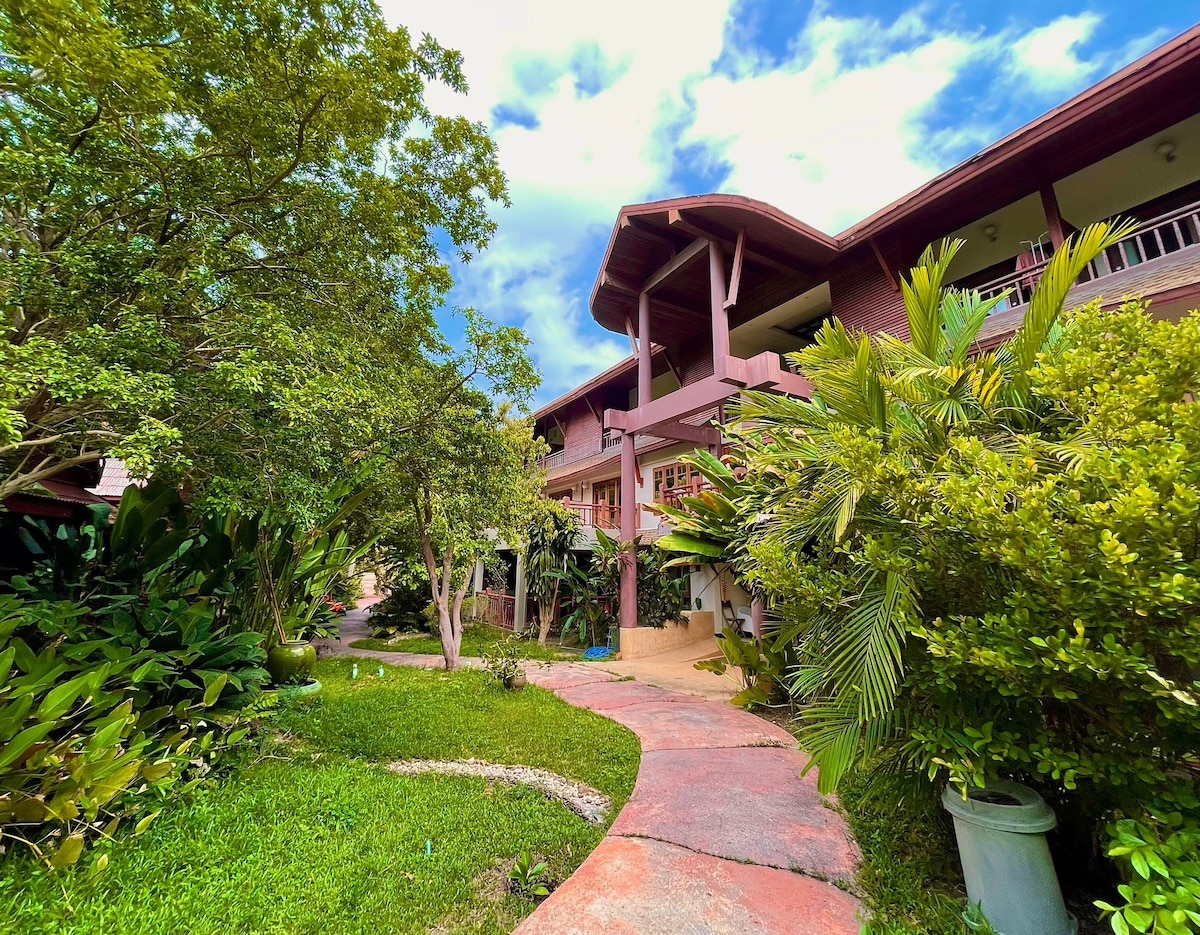 The exterior of the property is framed by lush greenery, featuring a winding path that leads to a multi-story structure. Natural light illuminates the vibrant foliage and tropical plants, creating a serene entrance to the building.