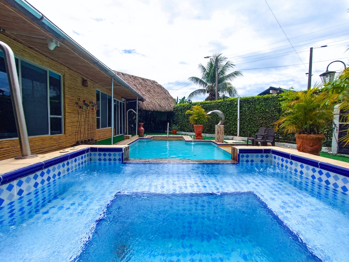 A spacious outdoor pool area is featured, showcasing a clear blue pool with a jacuzzi section. Surrounding greenery and palm trees provide a natural ambiance. A thatched-roof structure is visible in the background, enhancing the setting's relaxed atmosphere.