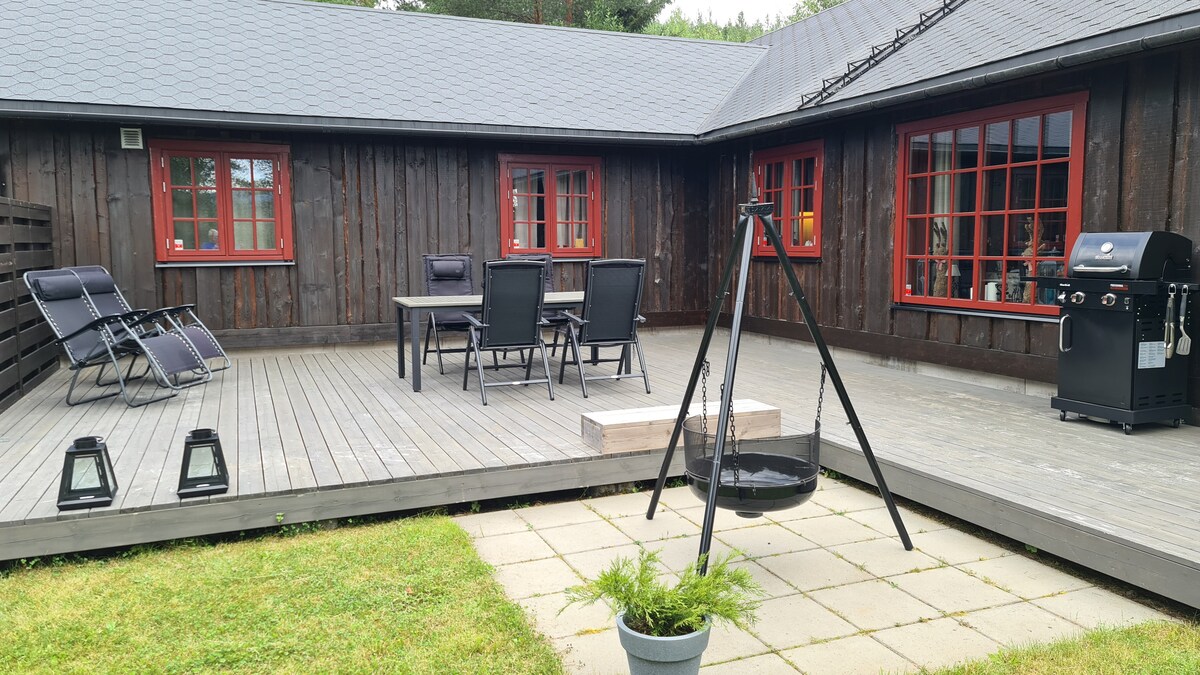 An outdoor deck is featured with comfortable seating for dining and relaxation. A gas grill is positioned nearby, and a fire pit adds a warm touch. Lanterns provide ambient lighting, while well-maintained greenery surrounds the patio area, creating an inviting outdoor space.