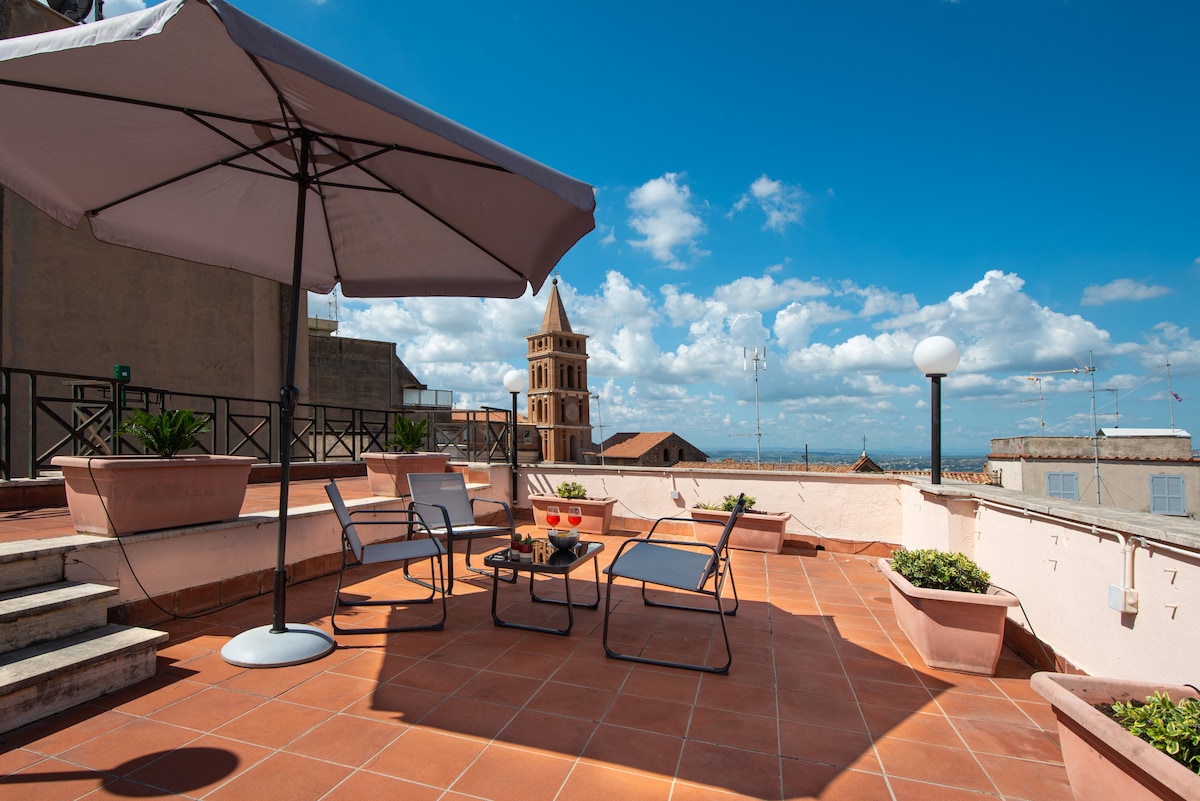 A spacious terrace is highlighted, featuring a large umbrella providing shade over four comfortable chairs. Potted plants add greenery to the space, complemented by terracotta tiles. In the background, a historic bell tower is visible against a bright blue sky with scattered clouds.