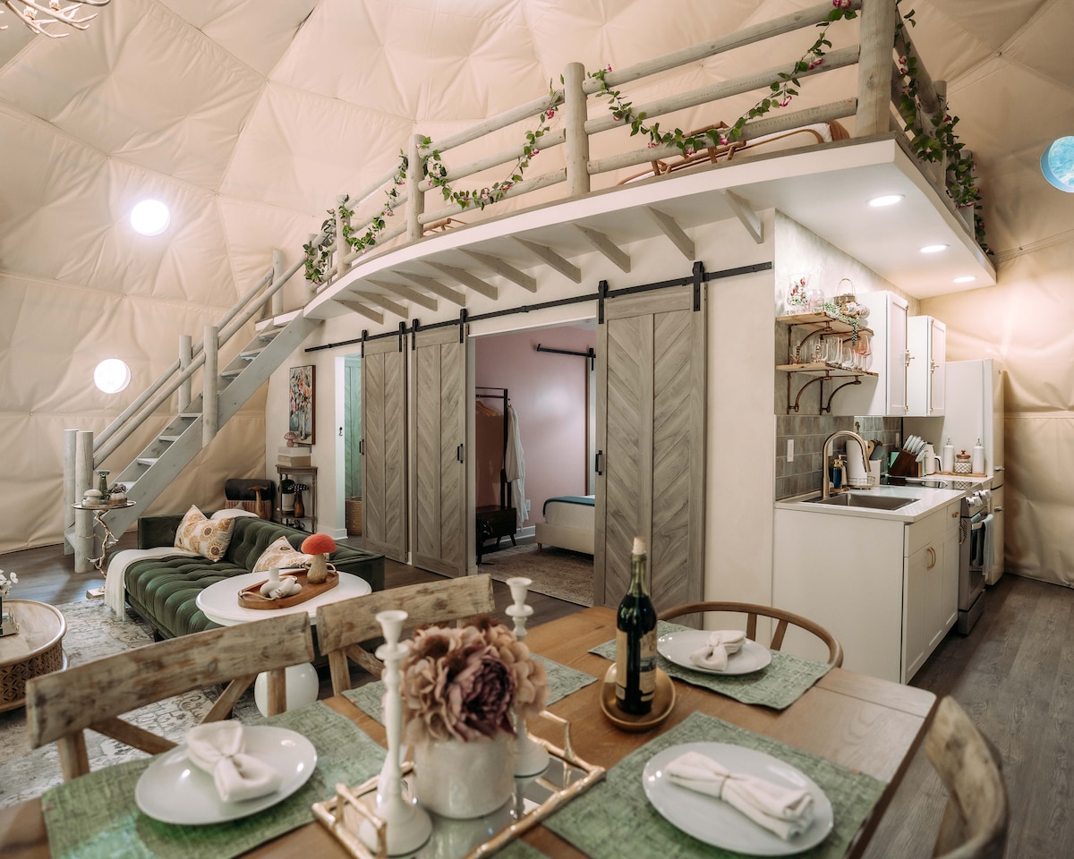 The interior space showcases an open-concept design with a spacious living area and a dining table set for six. Stylish barn doors lead to the bedrooms, while a loft area with wooden railing is visible above, accentuated by warm lighting and decorative elements.