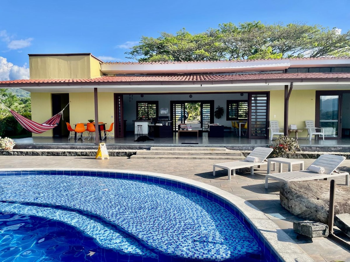 The exterior showcases a spacious pool area with a vibrant blue tile surface. Sun loungers are positioned nearby, complemented by bright orange chairs at a table. A cozy hammock is suspended, inviting relaxation, while the house features large sliding doors that connect to the outdoor space.