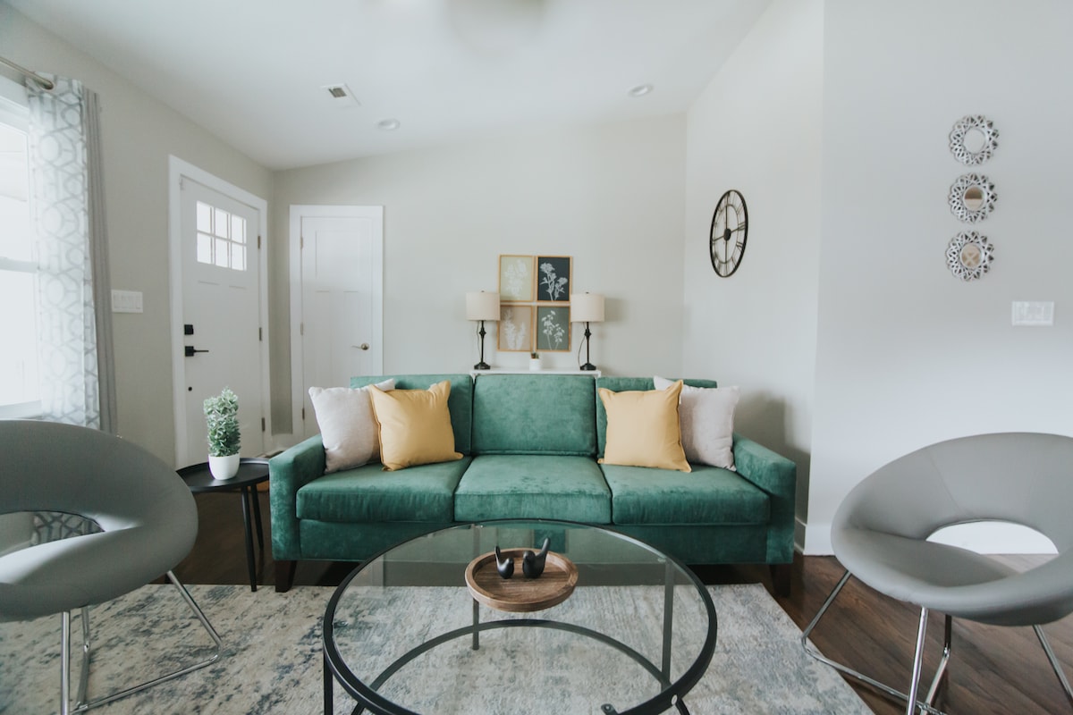 A cozy living room setup features a plush green sofa adorned with yellow pillows. A glass coffee table centers the space, complemented by modern chairs. Natural light filters through the windows, highlighting the minimal decor and neutral color palette.