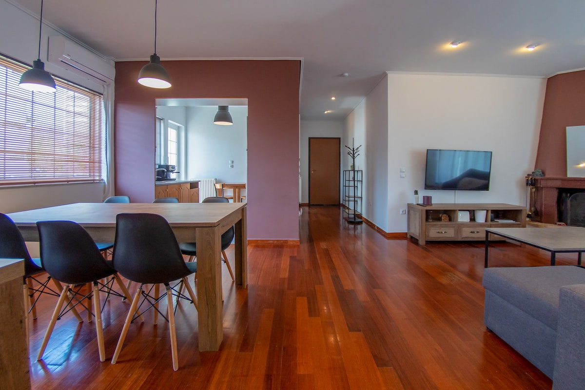 A spacious living area is highlighted by rich wooden flooring. A dining table, accompanied by several chairs, occupies one corner. A comfortable couch sits in front of a television, with natural light filtering through large windows, creating an inviting yet refined atmosphere.