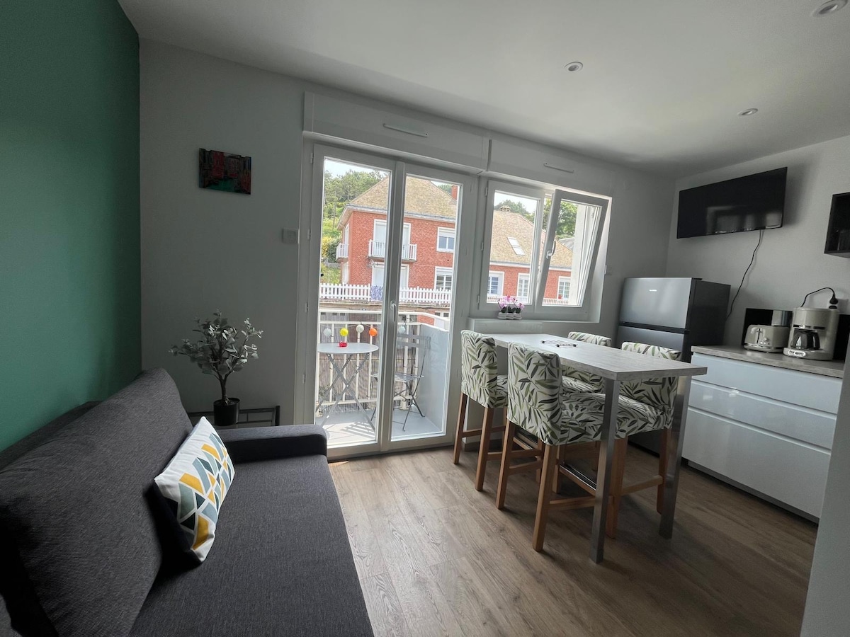 A modern living area is featured with a dark sofa positioned adjacent to large glass doors that open to a balcony. A small dining table with tropical-patterned chairs is visible, complementing a bright and airy kitchen space equipped with essential appliances.