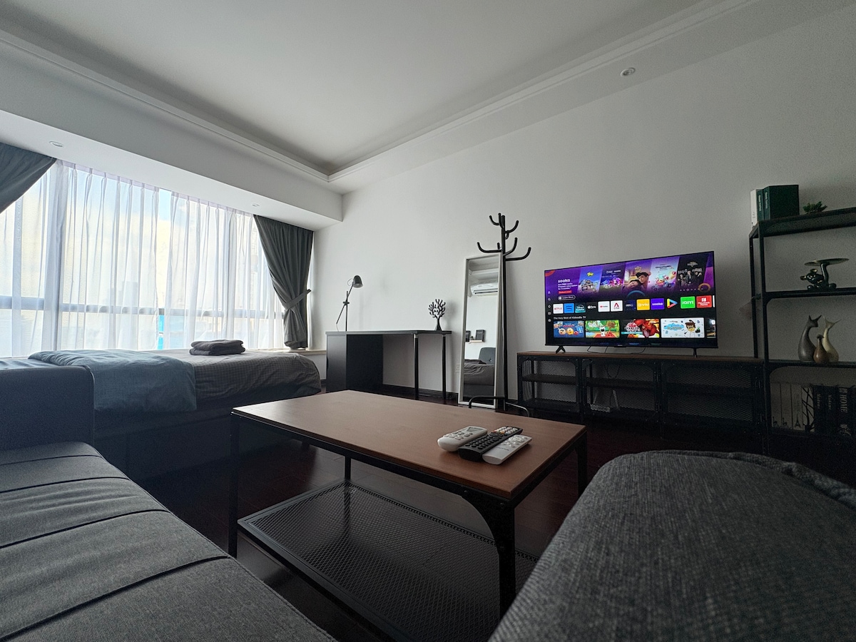 A spacious living area features large windows allowing natural light to fill the room. A comfortable bed is situated to one side, while a wooden coffee table stands in front of a sleek television. A mirror and a coat stand are positioned nearby.