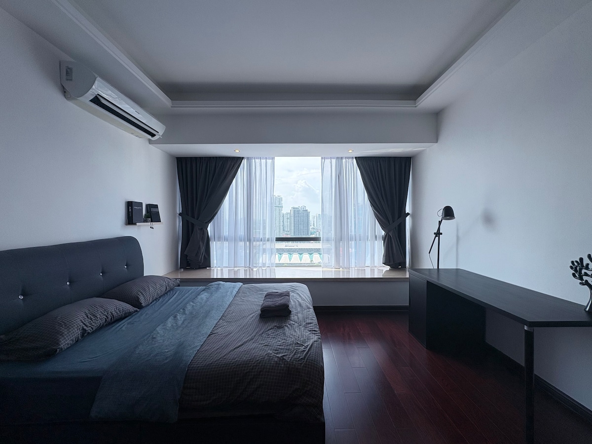 A spacious bedroom features a large bed with a dark bedding set. Floor-to-ceiling windows are dressed in light drapes, offering a view of the skyline. A sleek desk stands beside the window, and air conditioning units are mounted on the wall for comfort.