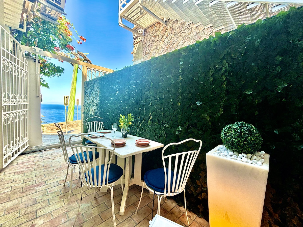 An outdoor terrace is presented with a small white dining table and four chairs, surrounded by a green hedge. A potted plant decorates one corner, while a scenic view of the sea is visible in the background, enhancing the inviting outdoor space.