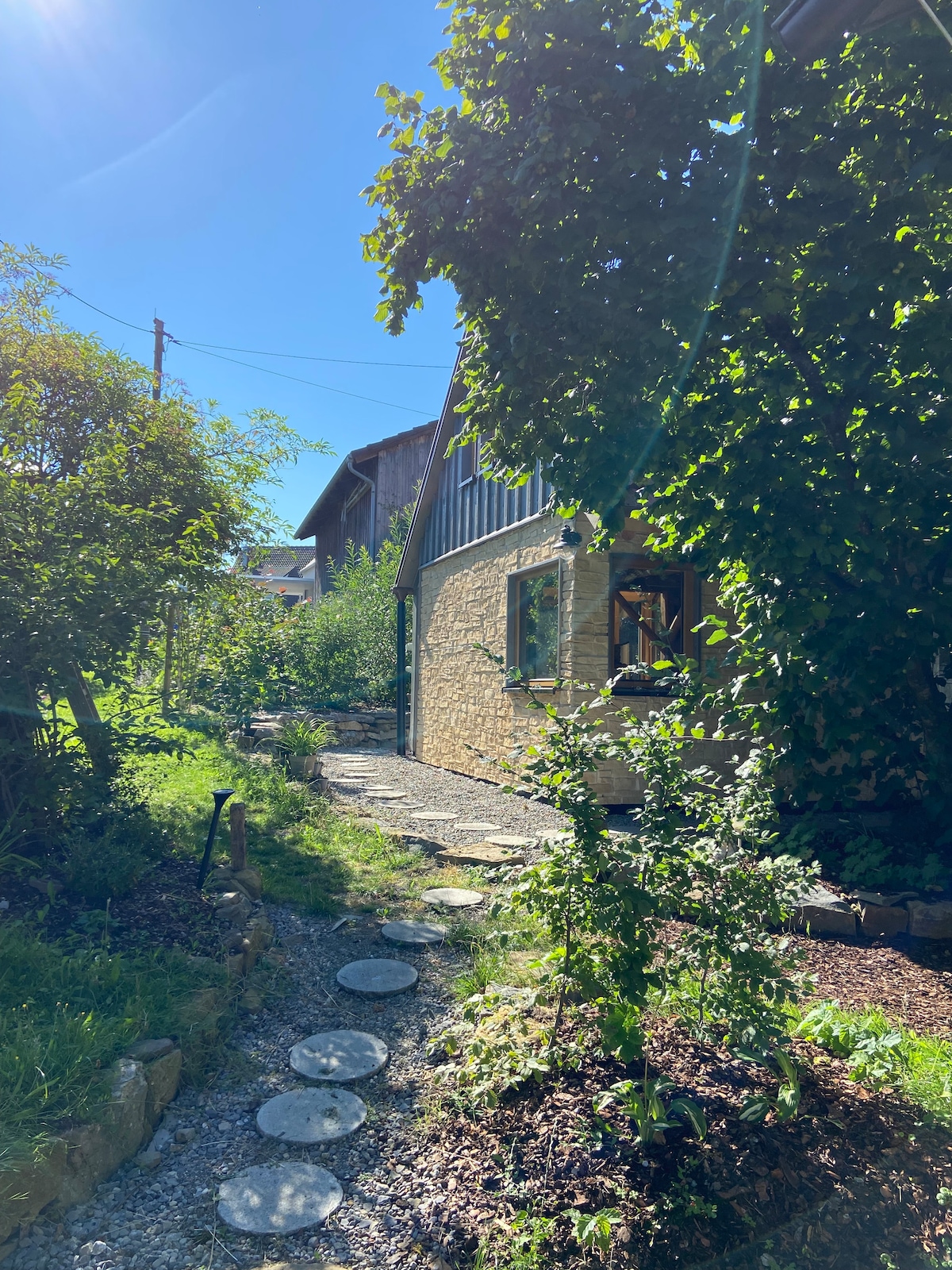 A charming pathway lined with natural stone slabs leads through lush greenery towards a warm-toned building. Sunlight filters through the leaves of nearby trees, creating dappled shadows on the ground, inviting guests to explore the peaceful surroundings.