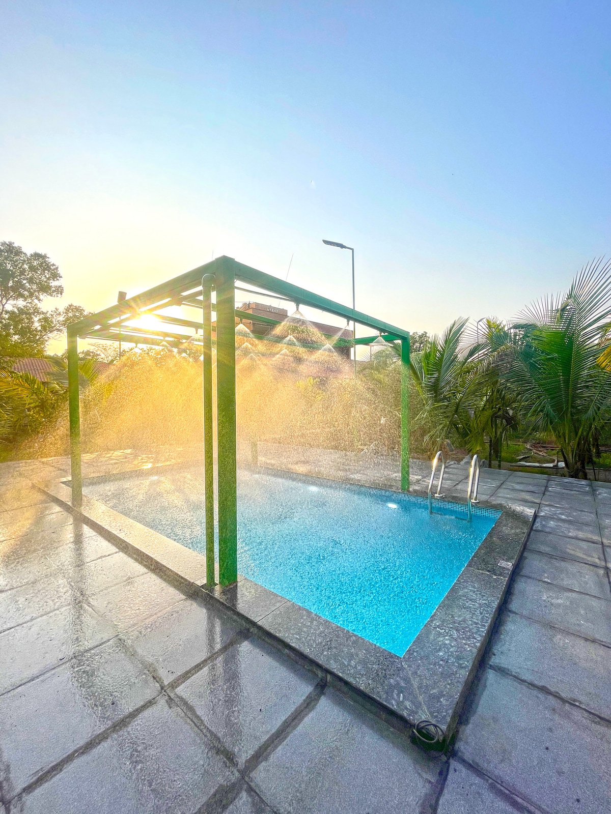 A refreshing swimming pool is shown, featuring a lush green canopy above. Water sprays from overhead, creating a misty atmosphere. The pool's clear blue water reflects the sunlight, surrounded by palm trees and a clean stone patio.