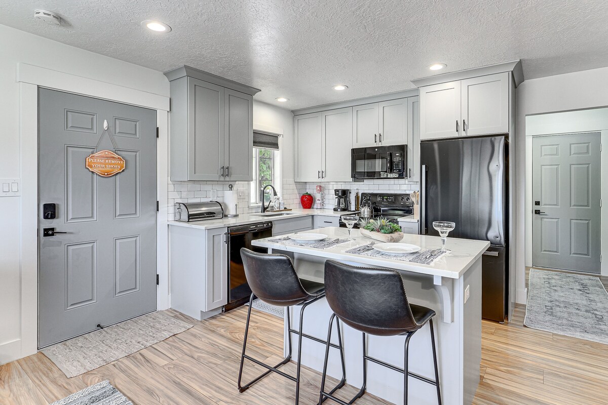 A modern kitchen features gray cabinetry and stainless steel appliances. A central island with two black bar stools is ready for dining, complemented by a light-colored countertop. Natural light enhances the space through a nearby window, creating a welcoming atmosphere.