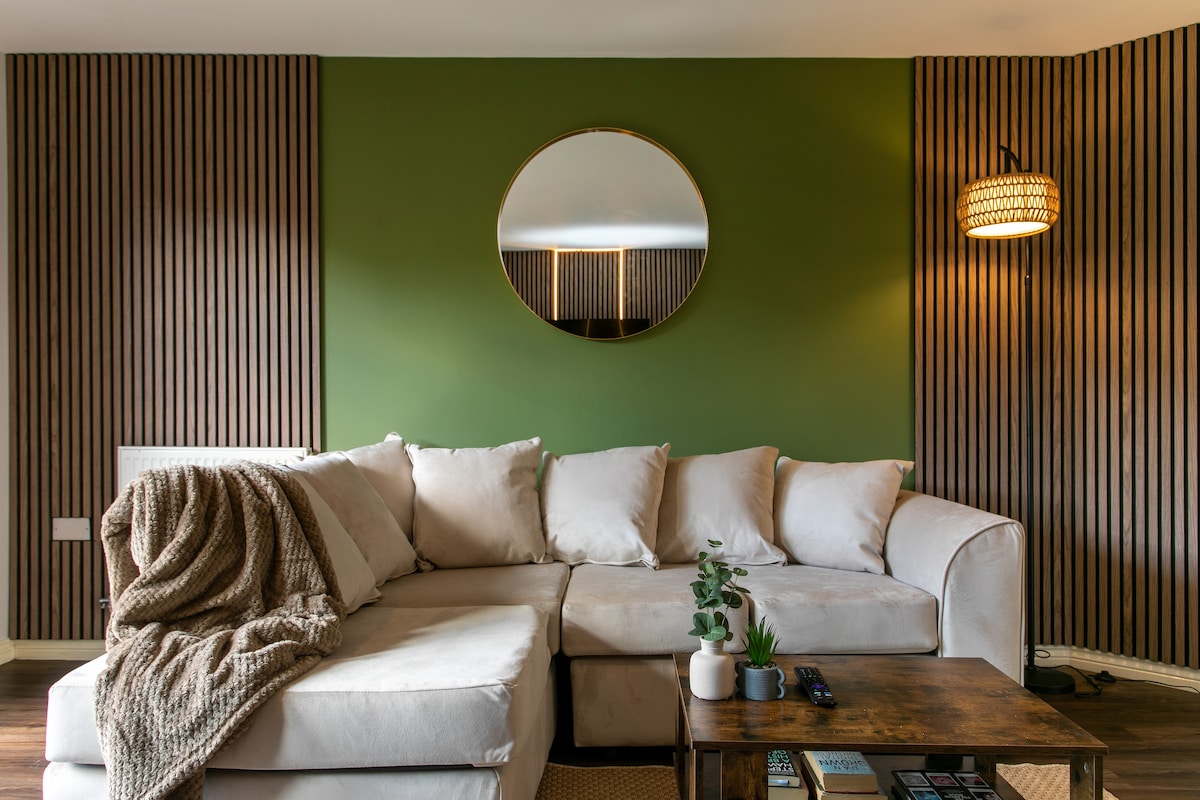 A cozy lounge area is highlighted by a sectional sofa adorned with soft throw blankets. A round mirror reflects the room’s warm colors, and a wooden coffee table sits in front, featuring small decorative plants and books. Soft lighting emanates from a stylish lamp.