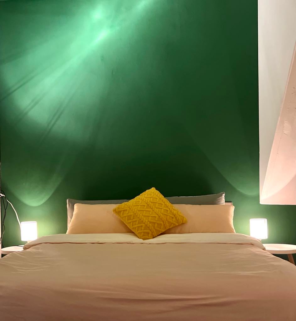 A spacious bed is presented with soft beige linens and a decorative yellow pillow. The green wall behind the bed is illuminated by gentle lighting, creating a relaxing ambiance. Two bedside lamps provide warm lighting, enhancing the overall tranquility of the space.