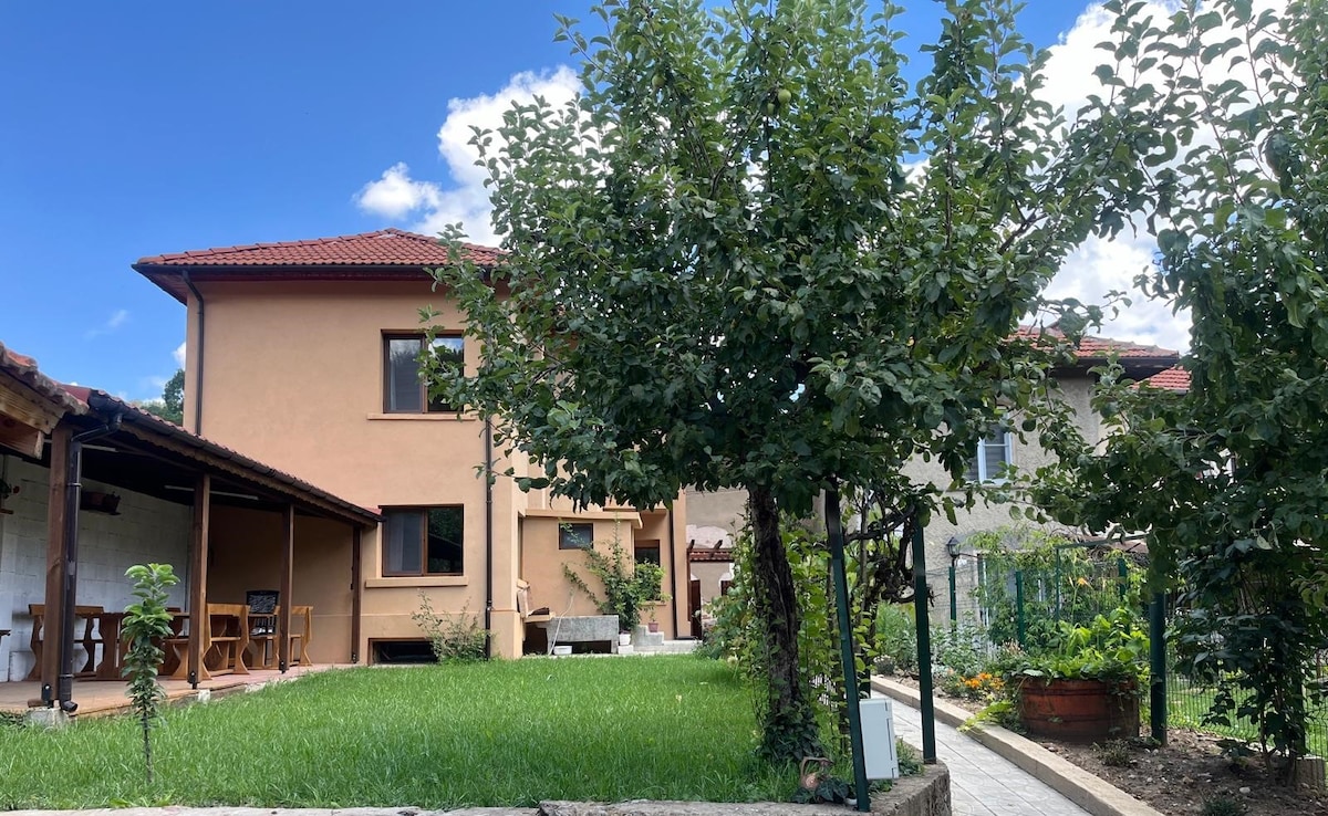 A spacious garden area is presented, featuring a well-maintained lawn and several trees. The exterior of the house is visible with warm-colored walls and a tiled roof. A covered porch is seen, furnished with a wooden table and chairs, inviting outdoor relaxation.
