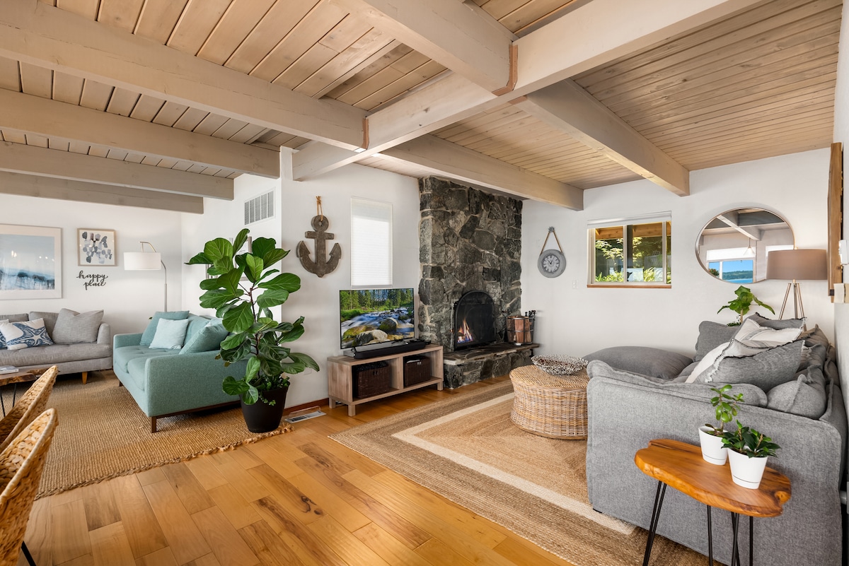 A spacious living room features a combination of comfortable seating options, including a sleek green couch and a cozy gray sofa. A television is placed on a wooden stand next to a fireplace framed by dark stones. Large windows allow light to fill the room, highlighting natural wood beams overhead.