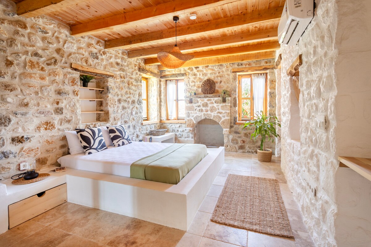 A spacious bedroom features stone walls and wooden beams, creating a natural ambiance. A large bed is positioned against a wall, complemented by decorative pillows. Two windows provide natural light, while a small potted plant adds a touch of greenery. A woven rug is placed on the stone floor.