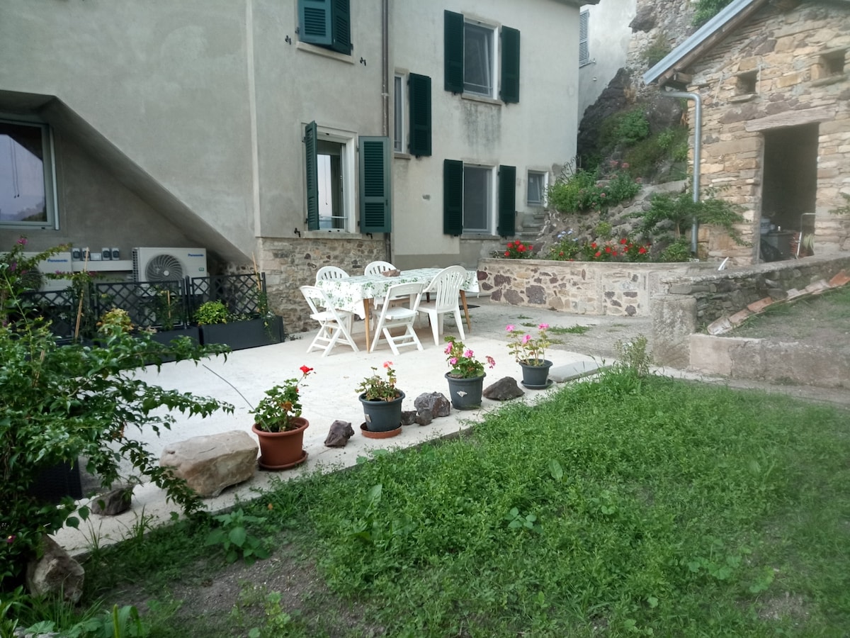 A shaded outdoor terrace features a sturdy table surrounded by four white chairs. Flower pots with colorful blooms line the edge, enhancing the natural setting. Stone walls and a grassy area provide a rustic backdrop, reflecting the charm of the countryside.
