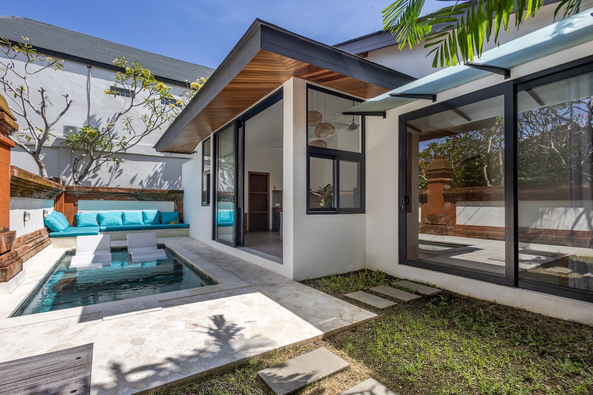 An outdoor area is highlighted by a compact pool, surrounded by lush greenery. A shaded seating area features teal cushions against a backdrop of large glass doors, creating an inviting space for relaxation. The clean lines of modern architecture are complemented by natural elements.