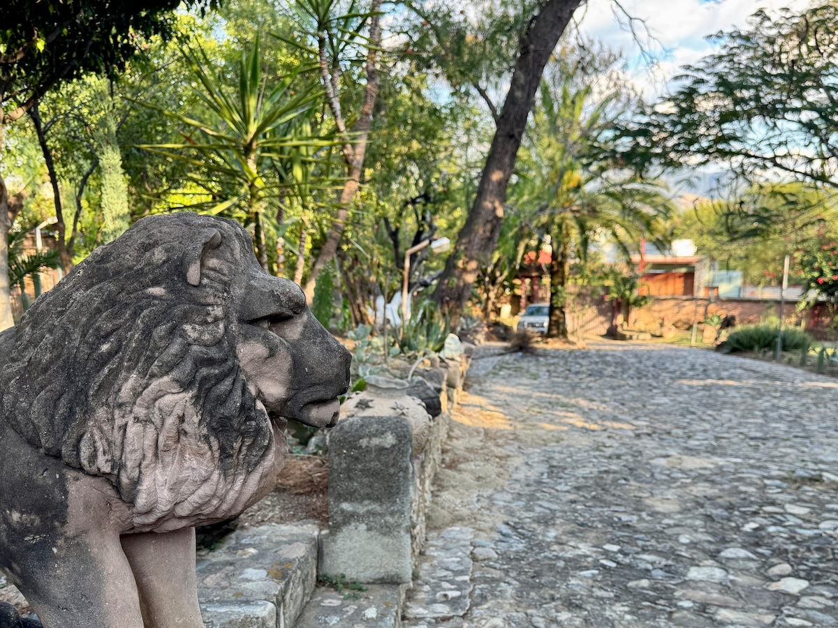 A stone lion statue is positioned on a stone pathway lined with greenery. Palms and other trees create a natural backdrop, while a distant structure is partially visible along the winding road, inviting exploration and tranquility.