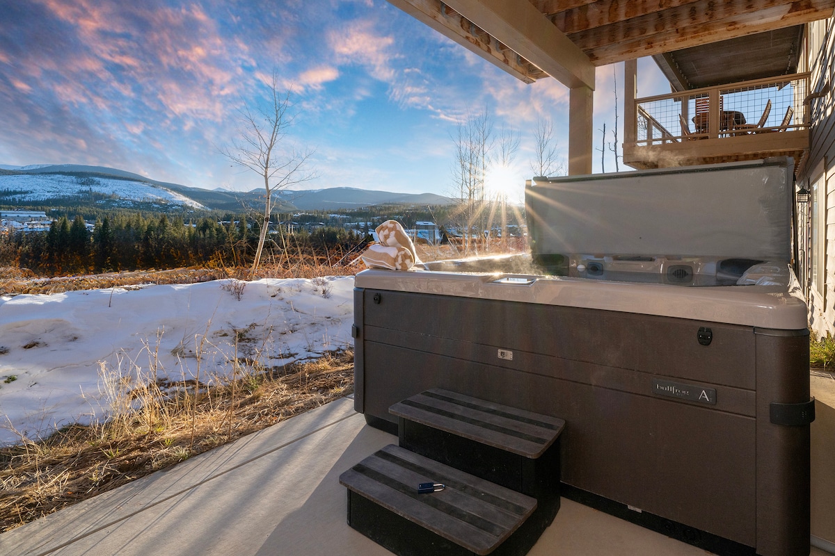 A spacious hot tub is positioned on a patio, offering views of the snow-covered landscape and distant mountains. Bright sunlight casts soft reflections on the water, while surrounding trees are visible under a colorful sky.