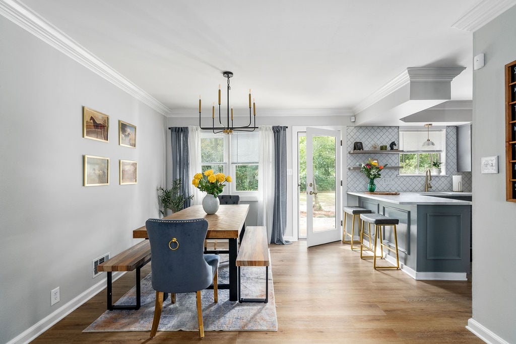 A dining area features a wooden table surrounded by upholstered chairs, illuminated by natural light from large windows. A modern kitchen bar with stylish stools is seen in the background, complemented by a decorative vase filled with fresh flowers.