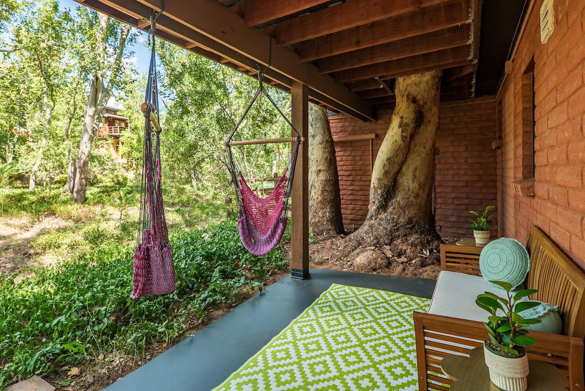 A shaded outdoor patio features a woven hammock and a hanging chair, surrounded by lush greenery. A wooden seating area is complemented by potted plants, with a patterned rug laid on the floor. Tall Sycamore trees provide natural shade, enhancing the serene setting.