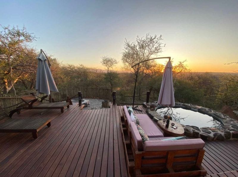 An outdoor deck is visible, featuring comfortable seating and a private plunge pool surrounded by natural stone. Sun loungers are positioned nearby, with trees and a serene landscape visible in the background, illuminated by the soft light of dawn.
