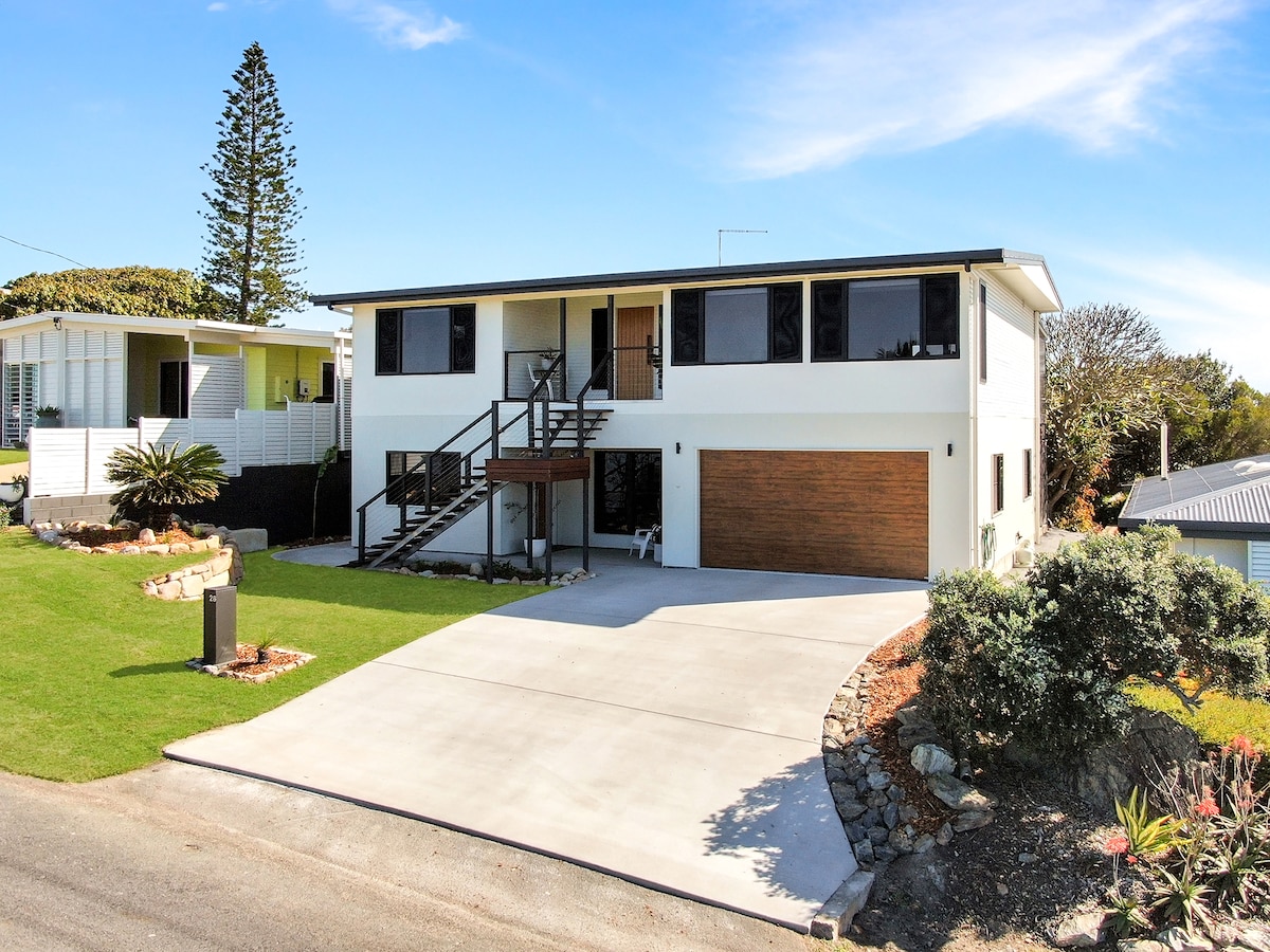 A modern two-level home is situated on a grassy lot. The façade features large windows and a welcoming front porch with steps leading to the main entrance. A spacious driveway provides access to a garage. Lush shrubs and landscaping enhance the curb appeal.