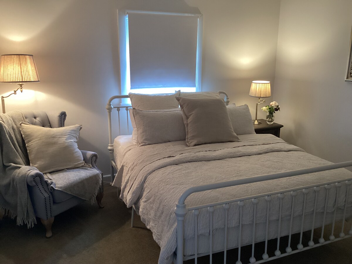 A soothing bedroom features a queen size bed dressed in soft linens, complemented by a decorative throw pillow. Two bedside lamps provide gentle illumination. A cozy armchair sits nearby, adorned with a plush blanket, offering a comfortable spot for relaxation.