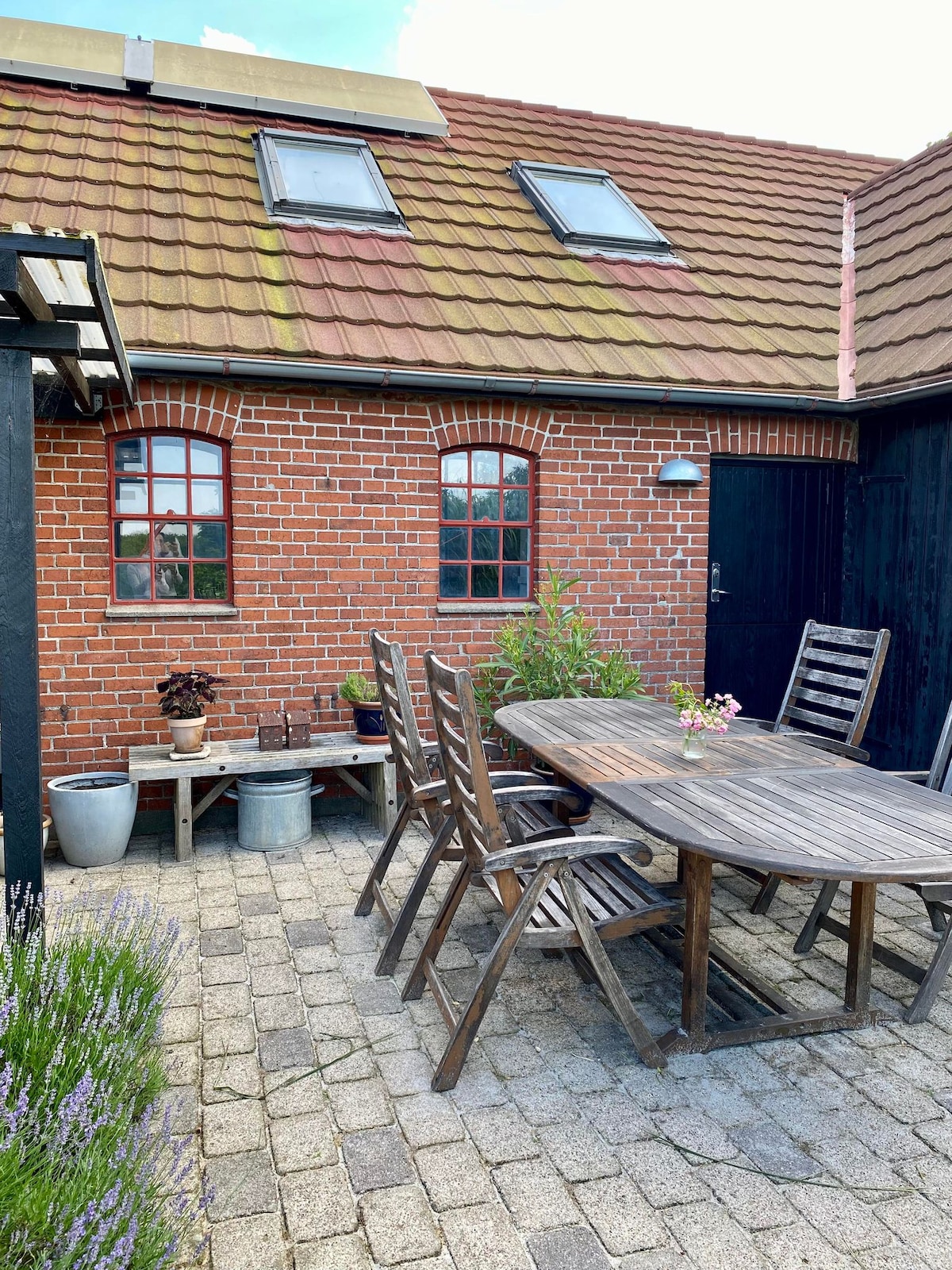 An inviting outdoor space features a wooden dining table surrounded by six chairs, set on a cobblestone patio. Potted plants and a small wooden bench add greenery, while an exterior wall of red brick with arched windows completes the rustic charm.
