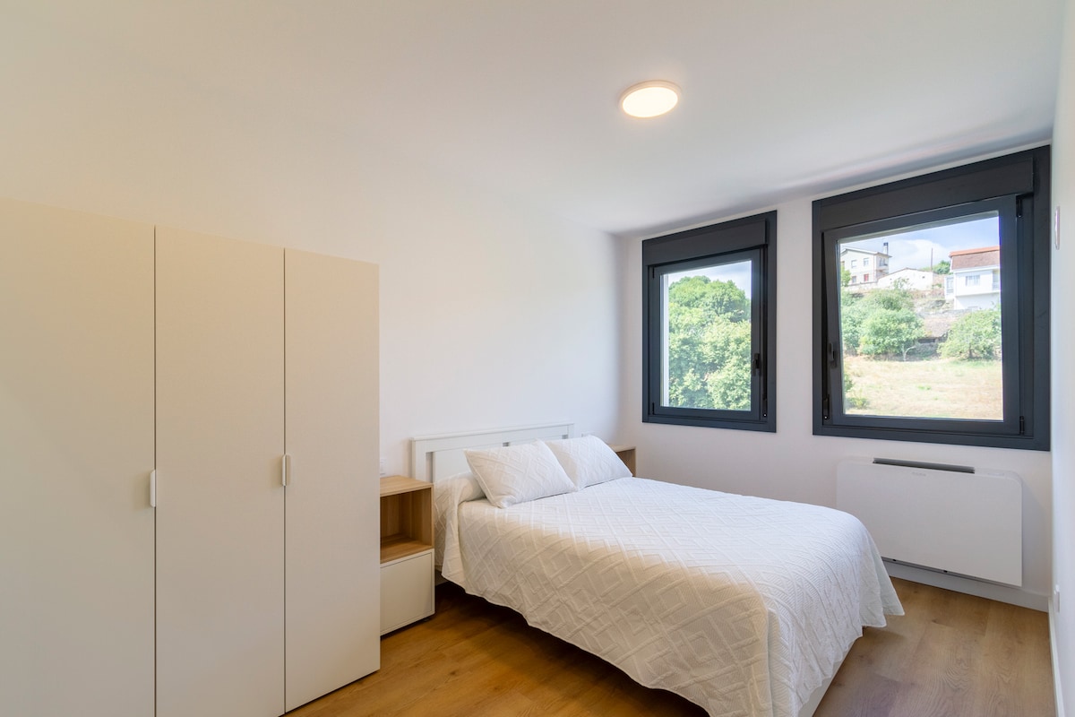 A light-filled bedroom features a spacious bed with a white quilt, complemented by a simple nightstand and a wardrobe. Two large windows provide a view of the surrounding greenery, allowing natural light to brighten the room.