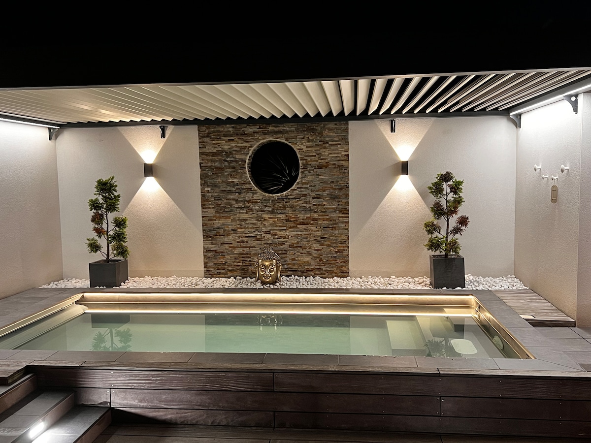 A private swimming pool is surrounded by wooden decking and accented by small white stones. A sleek stone wall provides a backdrop, illuminated by soft lighting on either side. Potted plants add a touch of greenery to the tranquil setting beneath a semi-covered pergola.