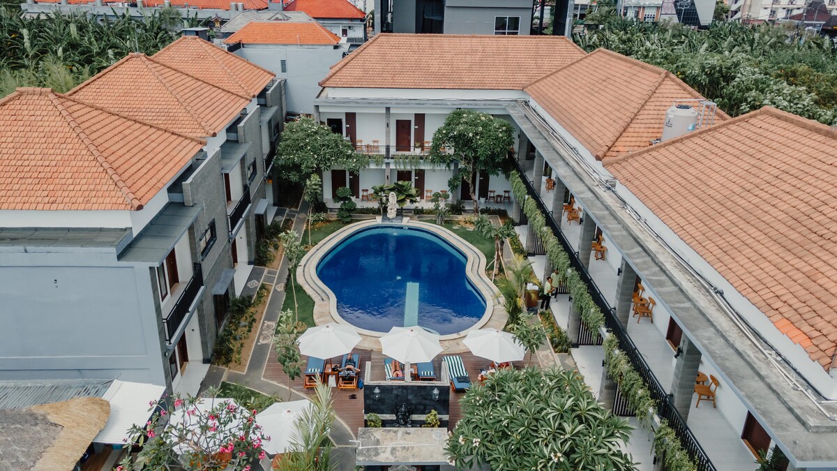 An aerial view captures the central outdoor swimming pool, surrounded by lush greenery and lounge chairs. The two-story buildings encircle the pool area, featuring terracotta roofs and balconies. Sun umbrellas provide shaded seating, creating a welcoming outdoor space.