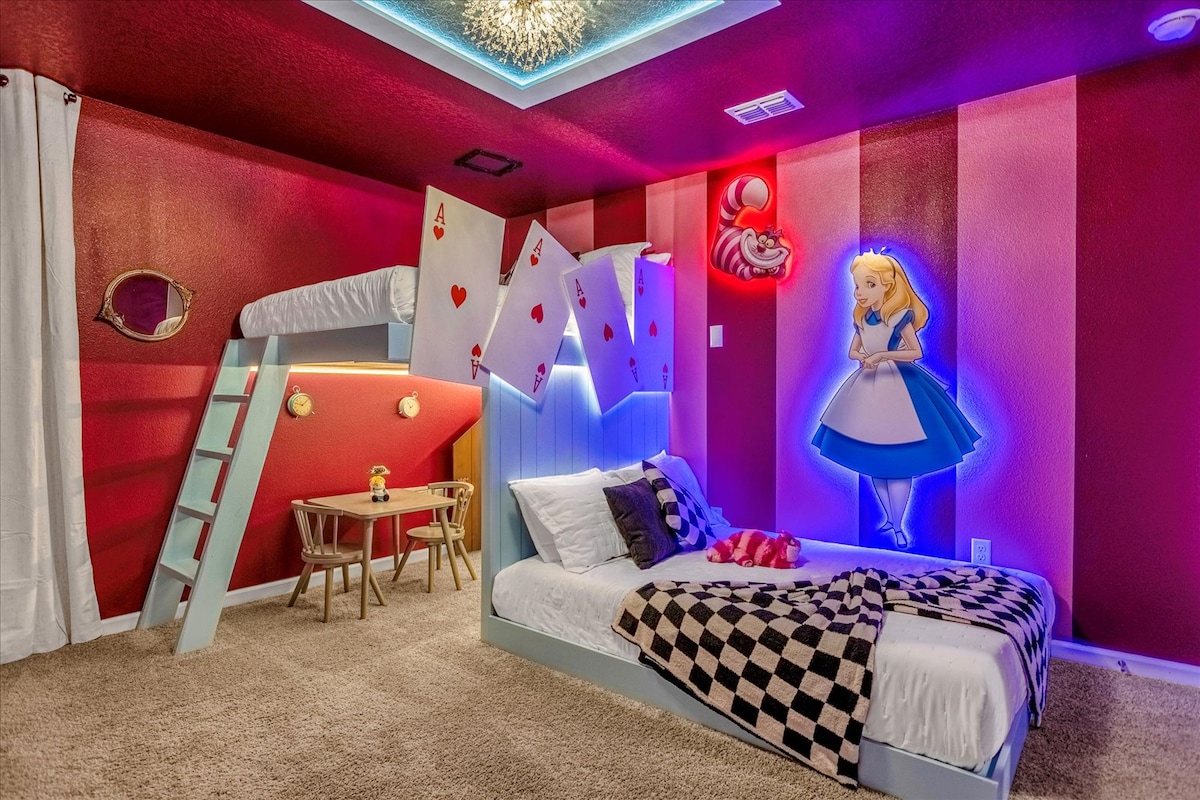 The themed bedroom is designed with vibrant red walls and whimsical décor. A lofted bed resembling playing cards stands above a cozy sleeping area. Colorful light fixtures illuminate the space, featuring a graphic of Alice in Wonderland against the wall. A small table and chairs complete the setting.