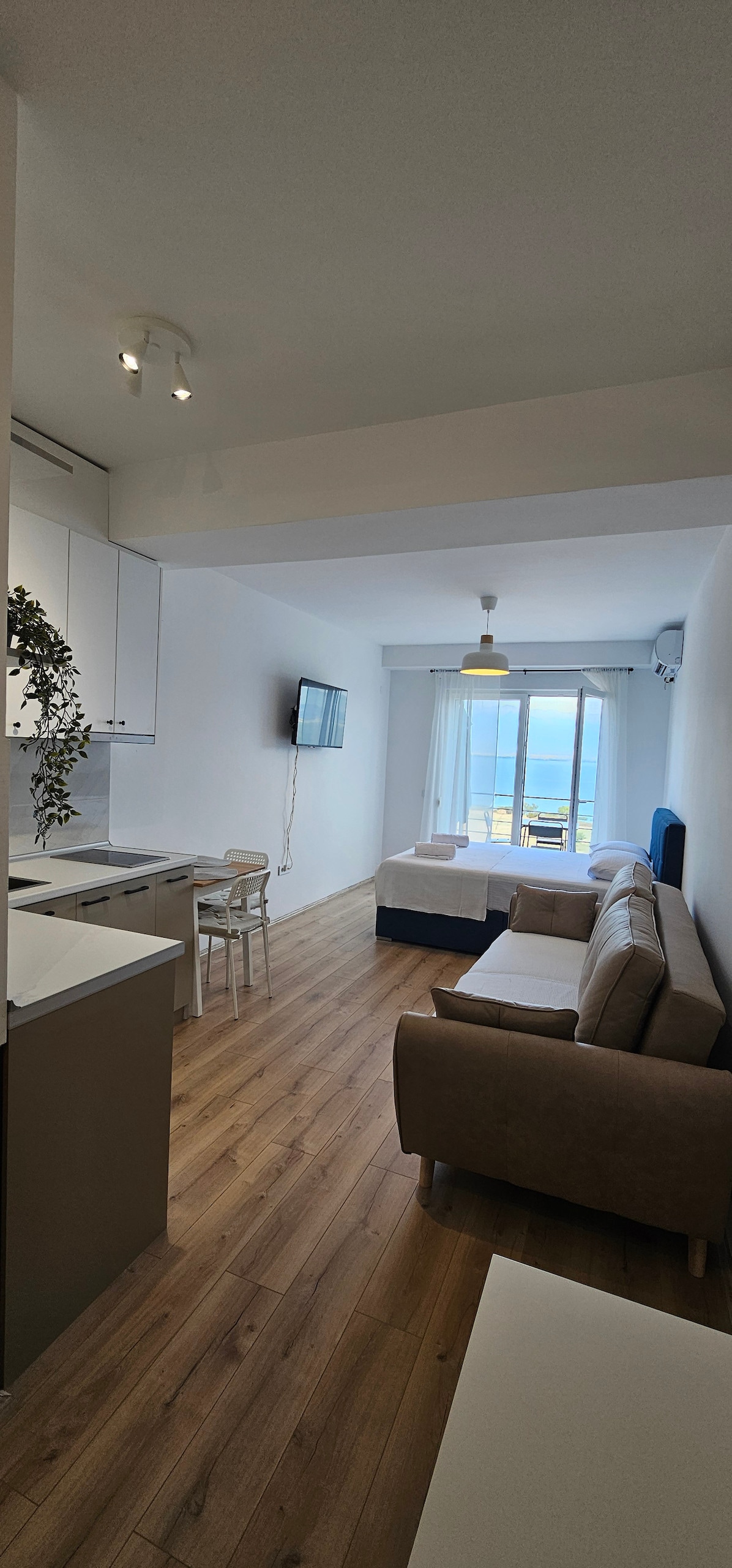 A bright and airy living space is displayed, featuring a cozy sofa facing a wall-mounted television. The kitchenette is visible to the left, with a dining table near the window. Light enters through glass doors that open to a breathtaking view of the lake.