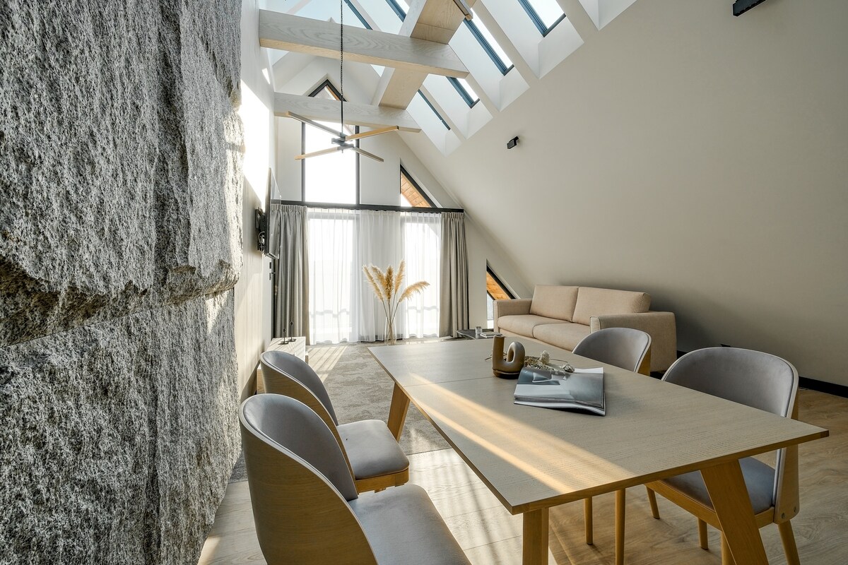 A bright living area is featured with vaulted ceilings and large skylights. A light wooden table surrounded by four chairs sits in the center, while a comfortable sofa is positioned against the textured stone wall. Sheer curtains softly diffuse light, enhancing the spacious feel.