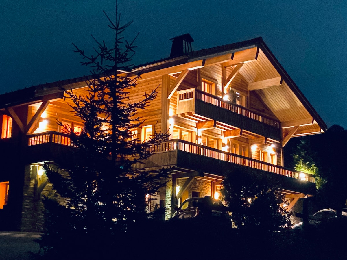 The chalet is illuminated at night, showcasing its wooden construction and architectural details. Balconies with wooden railings overlook the surrounding area. Soft lighting highlights the entrance and landscaping, creating an inviting ambiance.