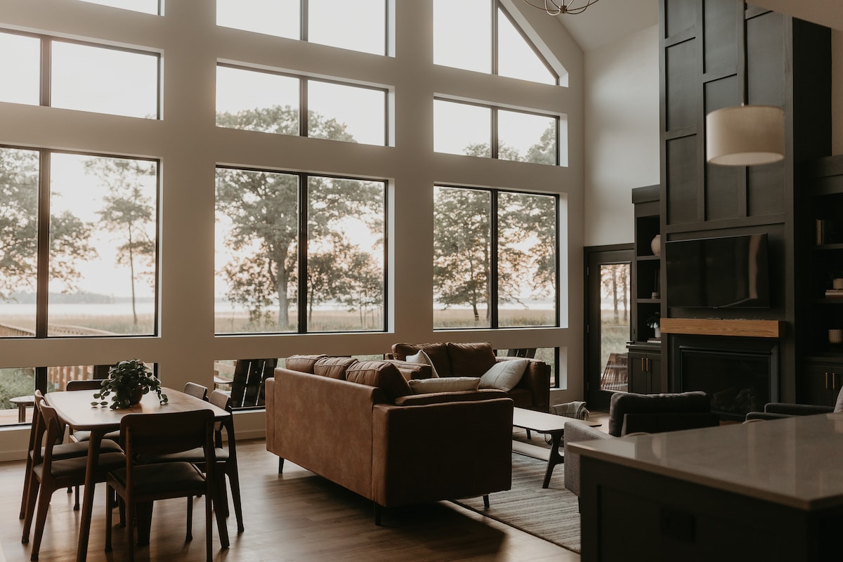 A spacious living area showcases large windows that frame views of surrounding trees and nature. The space features a comfortable couch, a dining table, and a modern entertainment unit. Natural light floods the room, creating a bright and airy atmosphere.