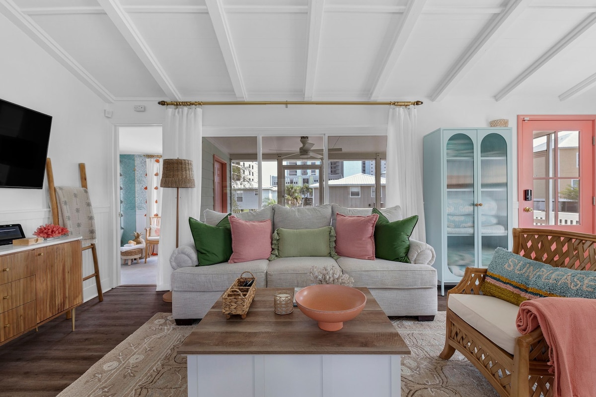 The living area features a comfortable seating arrangement with a plush sofa adorned with colorful cushions. A light wood coffee table sits at the center, paired with a decorative bowl. Large windows allow natural light to fill the room, showcasing a welcoming and airy atmosphere.