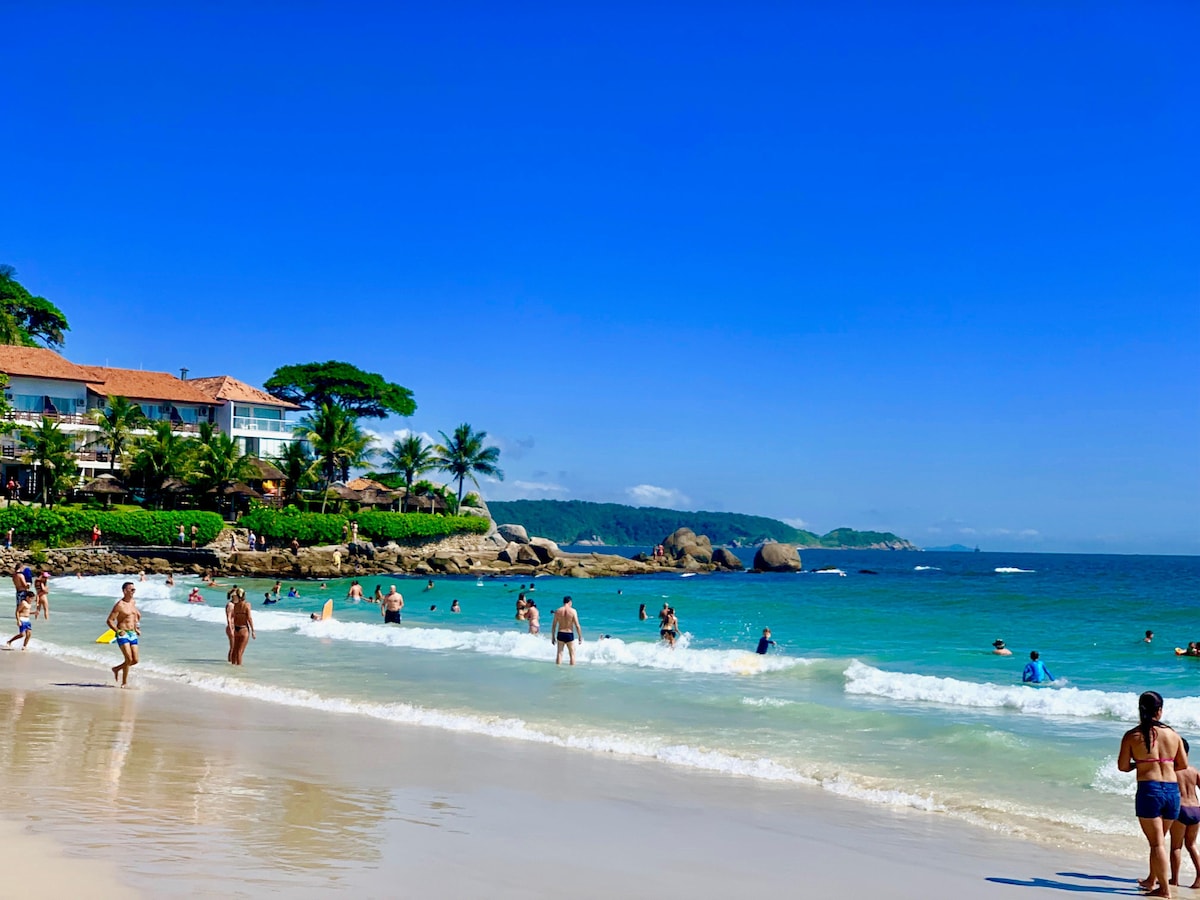 A vibrant beach scene is displayed, with individuals enjoying the water and soft waves lapping onto the sandy shore. Palm trees line the beach, and a multi-story building is visible in the background, adding to the coastal charm of this area.