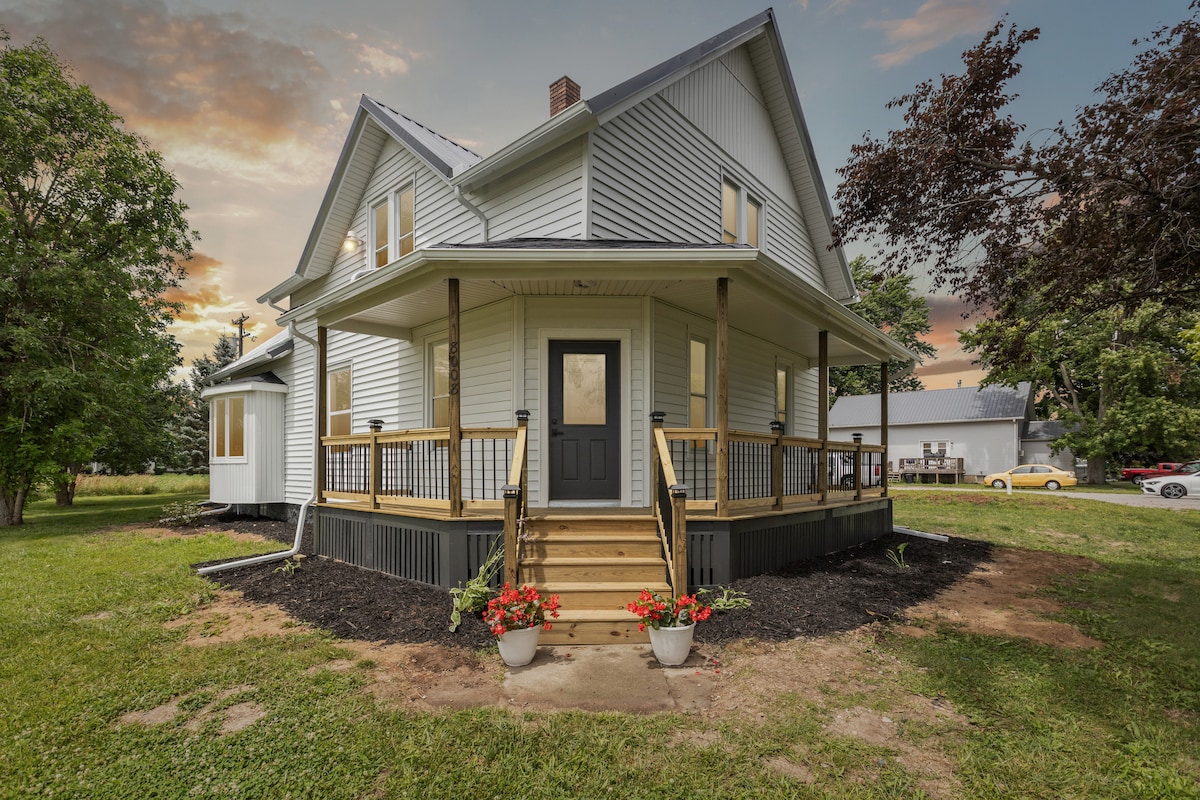 The exterior of a modern farmhouse is presented, featuring a welcoming front porch with wooden steps leading to a central door. Decorative planters with flowers enhance the space, while greenery surrounds the structure, creating a tranquil countryside setting.