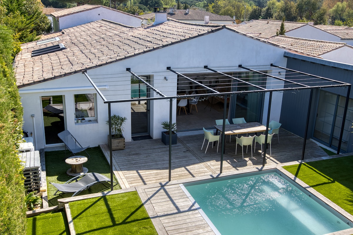 A spacious outdoor area features a wooden deck surrounding a swimming pool. Comfortable lounge chairs and a dining table with chairs are seen beneath a pergola. The villa's modern architecture is highlighted by large glass doors opening to the interior space.