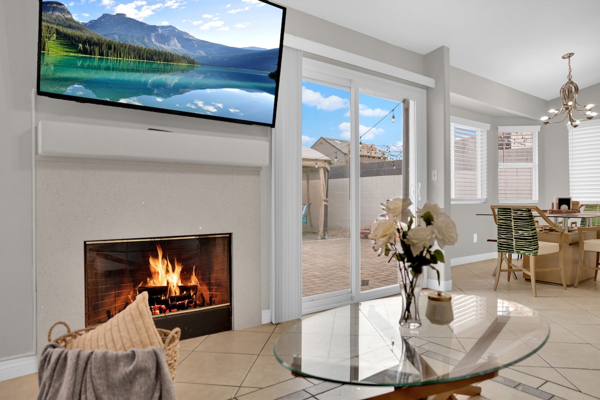 This inviting living area features a contemporary fireplace with a warm fire, a large wall-mounted TV displaying a scenic nature view, and a glass coffee table. Sliding doors open to a patio area, allowing natural light to fill the space.