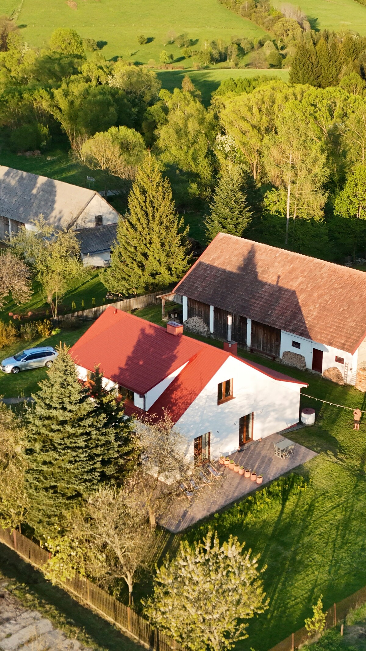 An aerial view presents a charming home with a red roof nestled among greenery. Surrounding trees and manicured lawns create a peaceful environment. Nearby structures can be seen, blending harmoniously into the natural landscape, enhancing the sense of seclusion and tranquility.