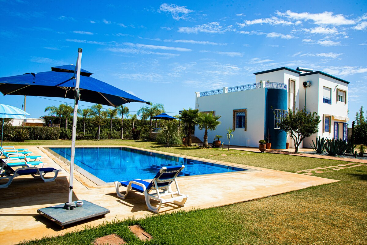 A spacious outdoor area features a large swimming pool surrounded by sun loungers and shaded by blue umbrellas. A two-story house is seen in the background, set against a clear blue sky and lush greenery.