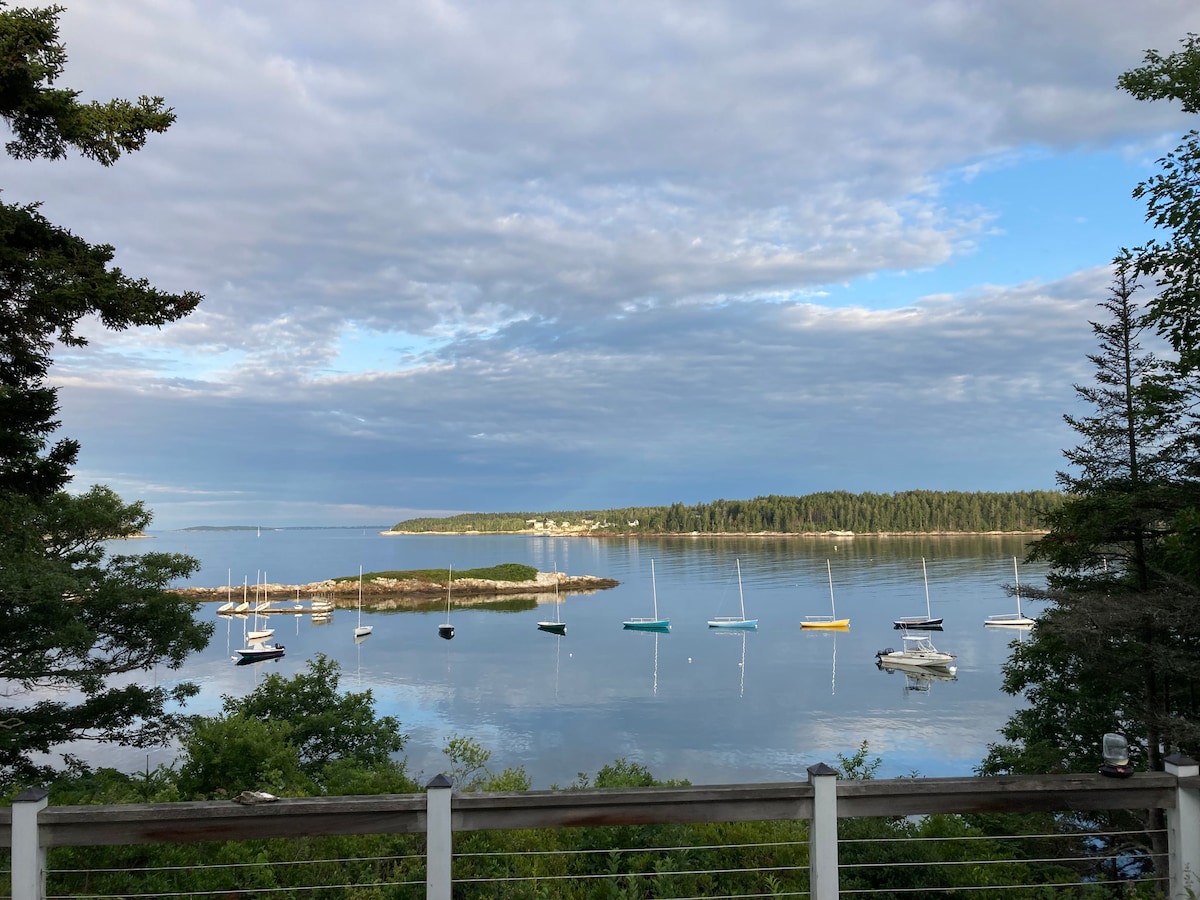 A tranquil view of Small Point Harbor is presented, featuring several sailboats gently anchored on calm waters. Lush greenery frames the scene, while scattered clouds drift across a bright sky, reflecting on the water's surface.