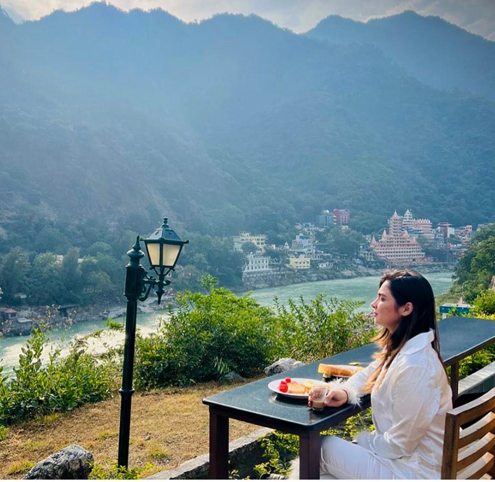 A dining table is set outdoors, overlooking a stunning view of the river and mountains. Lush greenery surrounds the area, complemented by a decorative lamp post. A plate with fresh fruits is placed on the table, inviting guests to enjoy a meal amidst nature.